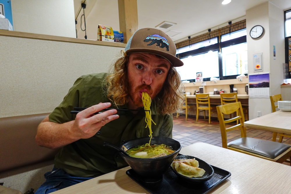 Samuel Jeffery comiendo ramen shio con gyoza en un restaurante de Hakodate, Hokkaido, Japón Samuel Jeffery comiendo fideos de un bowl de ramen shio en un restaurante informal de Hakodate, Hokkaido, Japón, con una porción de gyoza servida junto al ramen sobre la mesa.