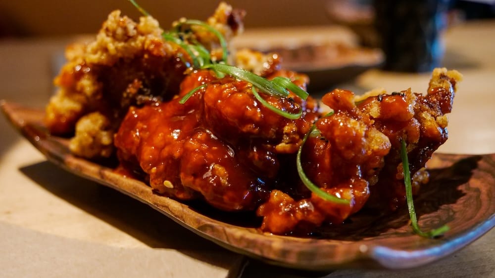 Pollo frito coreano con salsa picante en Shoku Izakaya, Banff, Alberta, Canadá Primer plano de pollo frito coreano en Shoku Izakaya, Banff, Alberta, Canadá, con trozos crocantes cubiertos por una salsa roja brillante y terminados con finas tiras de cebolla de verdeo sobre un plato de cerámica.
