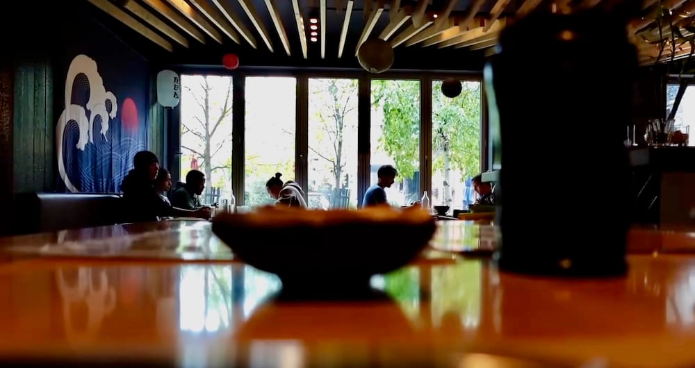Shoku Izakaya interior dining room, Banff, Alberta, Canada Interior dining room at Shoku Izakaya in Banff, Alberta, Canada, showing diners seated near large windows beneath a slatted wood ceiling and a wave-patterned mural, with a bowl in the foreground.