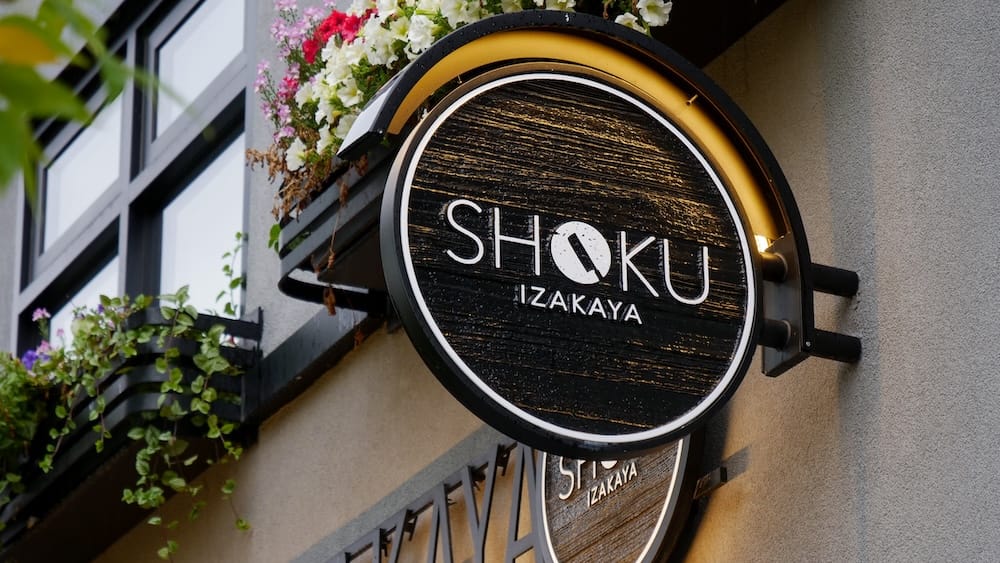 Shoku Izakaya restaurant sign, Banff, Alberta, Canada Round black and gold Shoku Izakaya restaurant sign mounted above a window with flower boxes on the exterior of the restaurant in Banff, Alberta, Canada.