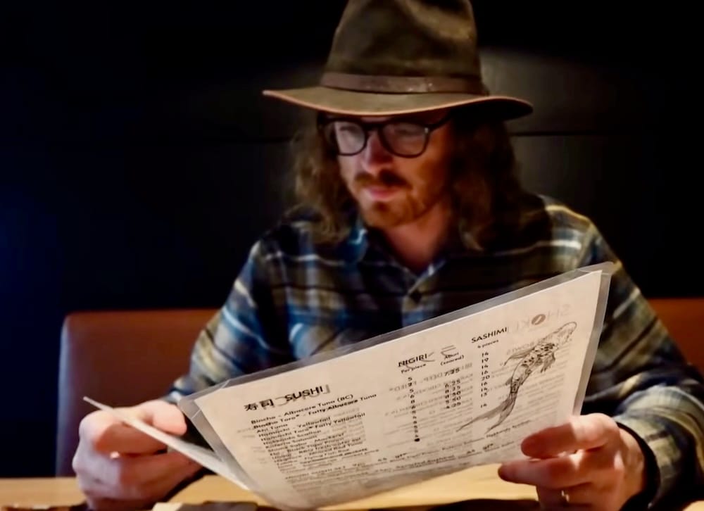 Samuel Jeffery reading the menu, Shoku Izakaya, Banff, Alberta, Canada Samuel Jeffery reads the sushi and sashimi section of the menu at Shoku Izakaya in Banff, Alberta, Canada, seated in a booth with the nigiri and sashimi price list visible.