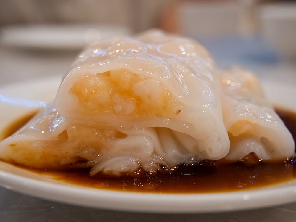 Close-up of a shrimp rice-crepe roll with visible shrimp filling, soaking in dark soy sauce on a plate at Chinese Cultural Centre Cuisine in Calgary, Alberta, Canada.