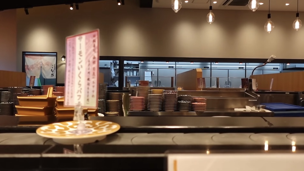 Cinta transportadora en Shunka Sushi (回転寿司 函館旬花), Goryokaku, Hakodate, Hokkaido, Japón, con cartel de menú y platos Cinta transportadora vacía en el restaurante de sushi Shunka en Hakodate, Hokkaido, Japón, con un cartel de menú en japonés de salmón e ikura sobre un soporte, pilas de platos y la cocina abierta detrás.