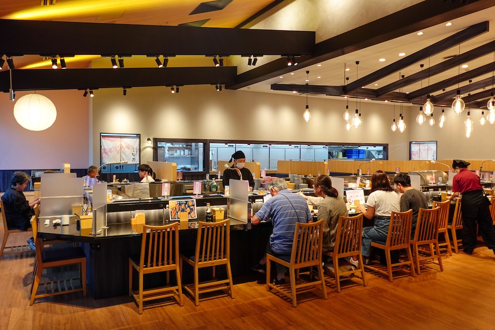 Salón comedor de Shunka Sushi (回転寿司 函館旬花), asientos en barra y cocina abierta, Goryokaku, Hakodate, Hokkaido, Japón Comensales sentados en sillas de madera a lo largo de una barra negra dentro del restaurante de sushi en cinta Shunka en Hakodate, Hokkaido, Japón, con chefs con barbijo en una cocina abierta bajo luces colgantes y vigas oscuras.