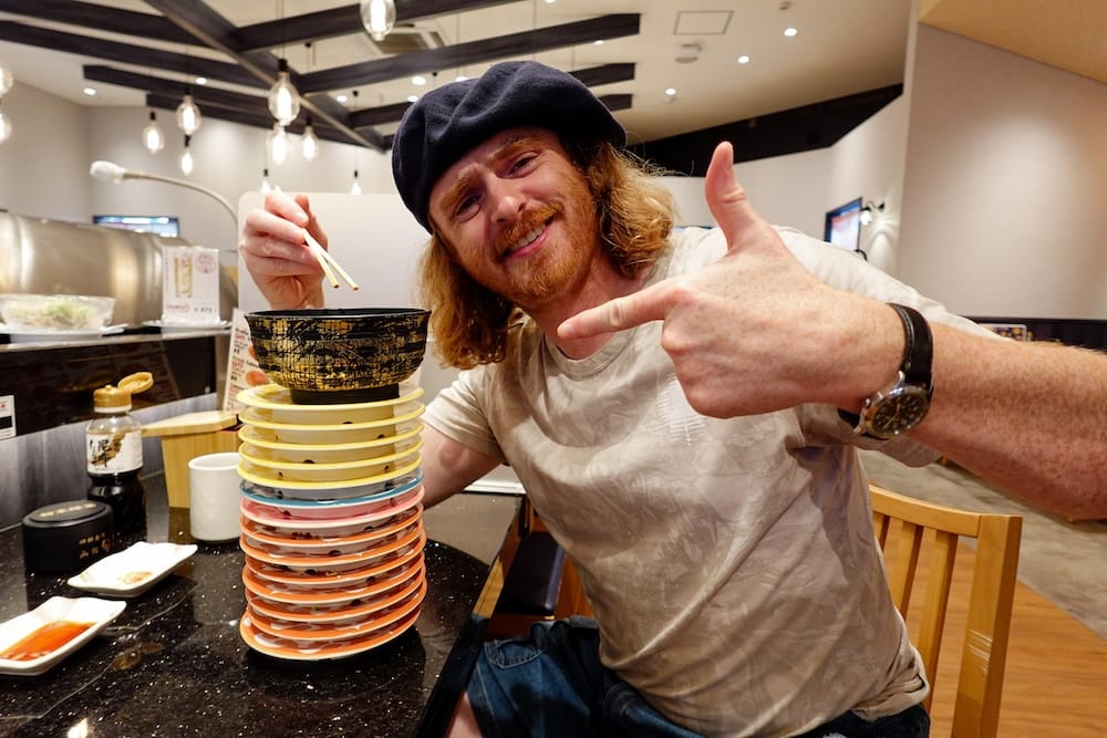 Samuel Jeffery, wearing a navy beret, smiles and points at a tall stack of empty plates topped with a bowl at Shunka Sushi, a conveyor-belt sushi restaurant in Hakodate, Hokkaido, Japan, chopsticks in hand.