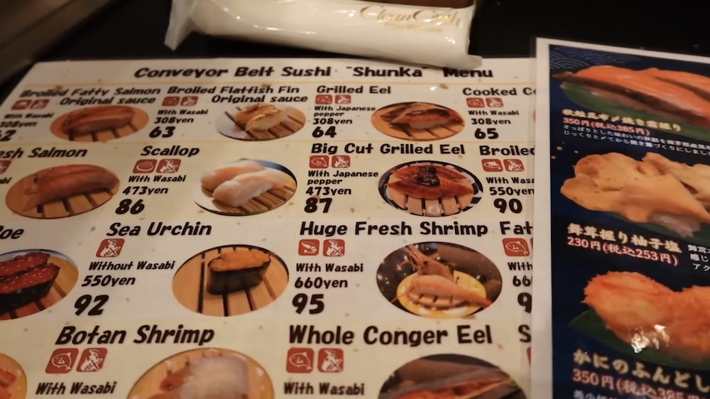 Menú en inglés con fotos y platos numerados, Shunka Sushi (回転寿司 函館旬花), Goryokaku, Hakodate, Hokkaido, Japón Menú plastificado con fotos en inglés de Conveyor Belt Sushi “Shunka” en Hakodate, Hokkaido, Japón, que muestra platos numerados como anguila grillada, vieira, erizo de mar y langostino, junto a una carta en japonés.
