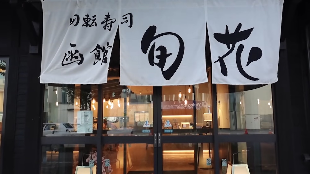 Cortina noren en la entrada de Kaiten-zushi Hakodate Shunka, Goryokaku-cho, cerca de la Torre Goryokaku, Hakodate, Japón Cortinas noren blancas con caligrafía negra que dice kaiten-zushi Hakodate Shunka (回転寿司 函館 旬花) cuelgan sobre la entrada vidriada del restaurante de sushi en cinta transportadora Shunka en Hakodate, Hokkaido, Japón.