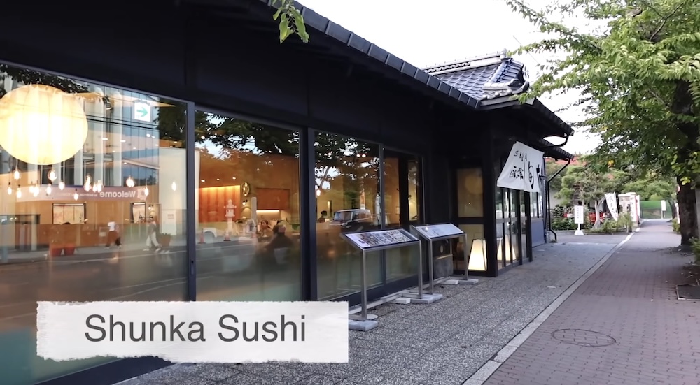 Fachada exterior de Shunka Sushi (回転寿司 函館旬花), edificio independiente en Goryokaku-cho, Hakodate, Hokkaido, Japón Exterior del restaurante de sushi en cinta transportadora Shunka en Hakodate, Hokkaido, Japón: un edificio oscuro con techo de tejas, grandes ventanales, cortinas noren en la puerta y carteles sobre una calle adoquinada.