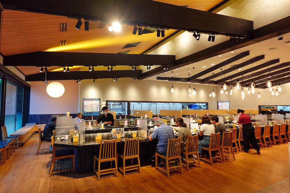 Interior del restaurante de sushi con cinta transportadora Shunka en Hakodate, Hokkaido, Japón, con una larga barra curva de sushi, sillas de madera, comensales sentados, personal preparando comida, vigas oscuras a la vista y luces colgantes cálidas.