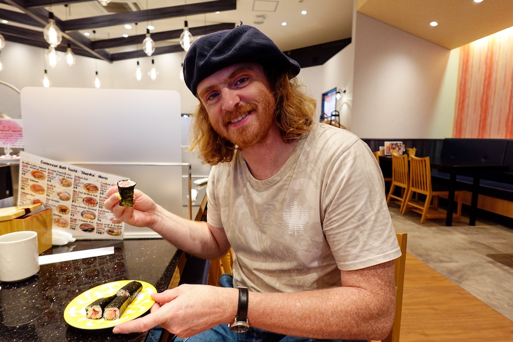 Samuel Jeffery disfrutando rolls maki en Kaiten Sushi Hakodate Shunka, restaurante de sushi con cinta transportadora en Hokkaido, Japón Samuel Jeffery sonríe mientras sostiene un roll de sushi envuelto en alga nori y un plato amarillo con piezas de maki en el restaurante de sushi con cinta transportadora Shunka, en Hakodate, Hokkaido, Japón, con un menú ilustrado detrás.