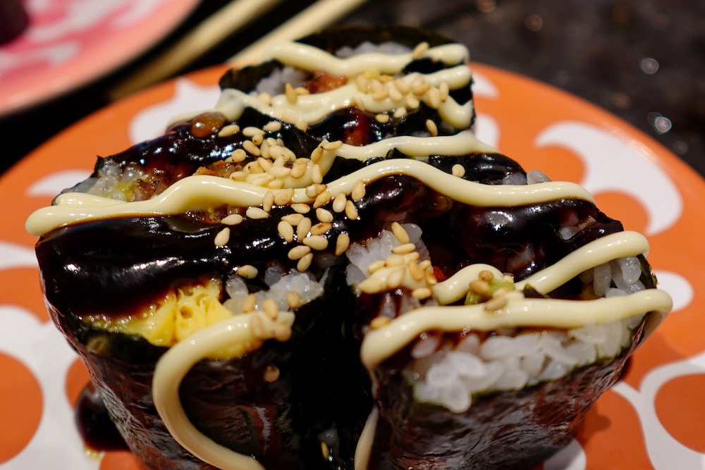 Primer plano de gruesos rolls de sushi en el restaurante de cinta transportadora Shunka en Hakodate, Hokkaido, Japón, envueltos en alga nori oscura con arroz y huevo visibles, cubiertos con salsa marrón brillante, mayonesa y semillas de sésamo en un plato naranja.