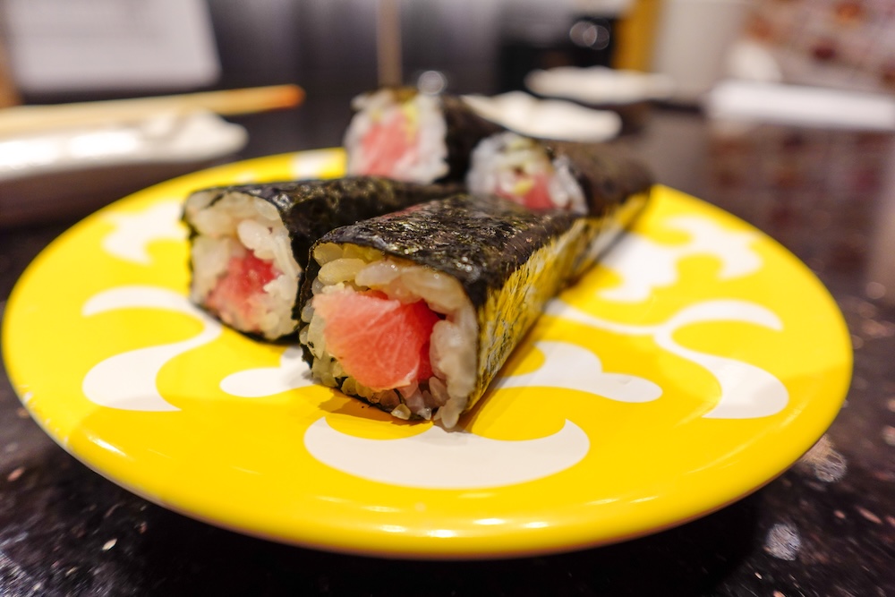 Primer plano de cuatro piezas de maki sushi de atún en el restaurante de cinta transportadora Shunka en Hakodate, Hokkaido, Japón, mostrando atún rosado y arroz blanco envueltos en alga nori oscura sobre un plato amarillo brillante.