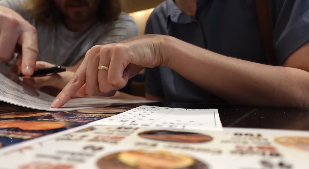 Eligiendo del menú en Shunka Sushi, Goryokaku, Hakodate, Hokkaido, Japón, con Audrey Bergner y Samuel Jeffery Audrey Bergner señala un menú con fotos mientras Samuel Jeffery sostiene una lapicera junto a un papel de pedido en blanco sobre la mesa del restaurante de sushi en cinta Shunka en Hakodate, Hokkaido, Japón.