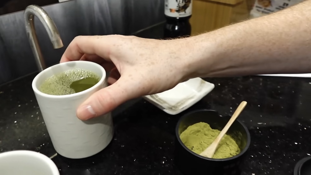 Matcha de autoservicio en la barra, Shunka Sushi (回転寿司 函館旬花), Goryokaku, Hakodate, Hokkaido, Japón, con Samuel Jeffery La mano de Samuel Jeffery sostiene una taza blanca de té verde espumoso junto a una lata de matcha en polvo con cuchara de madera y una canilla de agua caliente en el restaurante Shunka en Hakodate, Hokkaido, Japón.