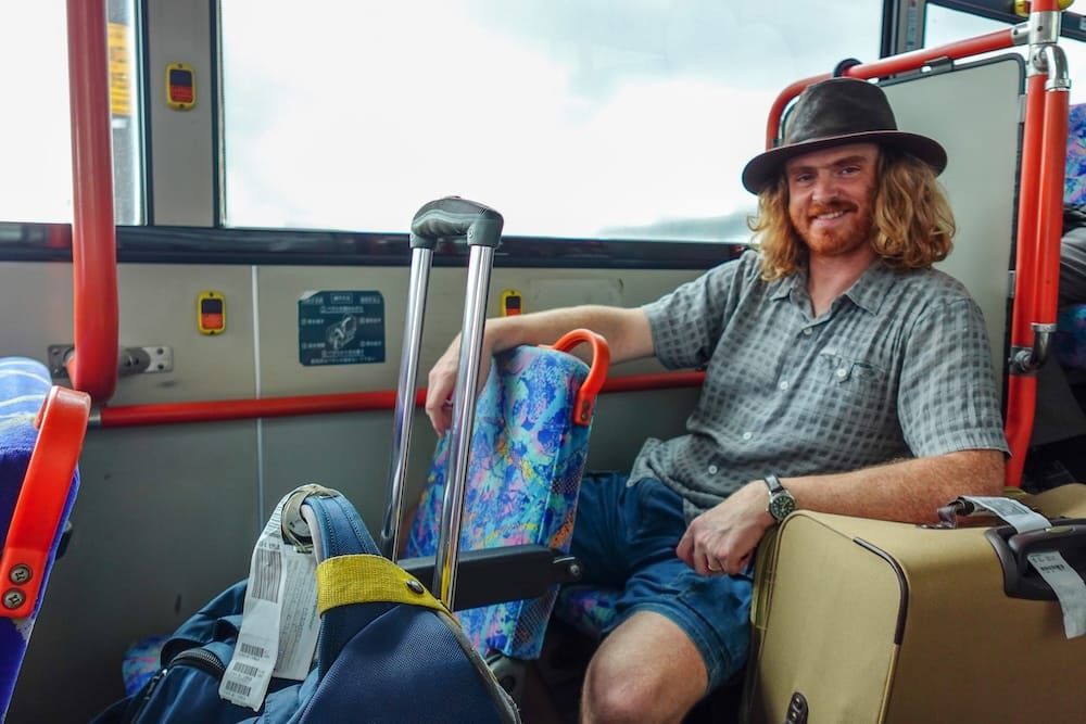 Arriving by shuttle bus with luggage, Hakodate, Hokkaido, Japan – Samuel Jeffery Samuel Jeffery sits on a shuttle bus surrounded by luggage on arrival in Hakodate, Hokkaido, Japan, wearing a hat and holding onto a rolling suitcase beside a large checked bag.