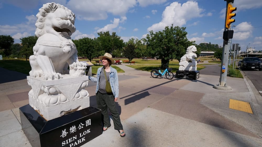 Audrey Bergner junto a un león guardián chino blanco en Sien Lok Park, Calgary, Alberta, Canadá, con una segunda estatua, senderos, árboles e infraestructura urbana al fondo.