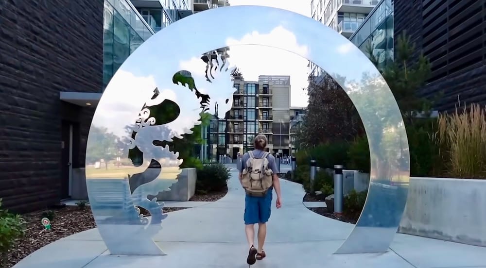 Samuel Jeffery camina a través de una gran instalación circular de metal cerca de Sien Lok Park, en el centro de Calgary, Alberta, Canadá, rodeado de jardines y modernos edificios residenciales.