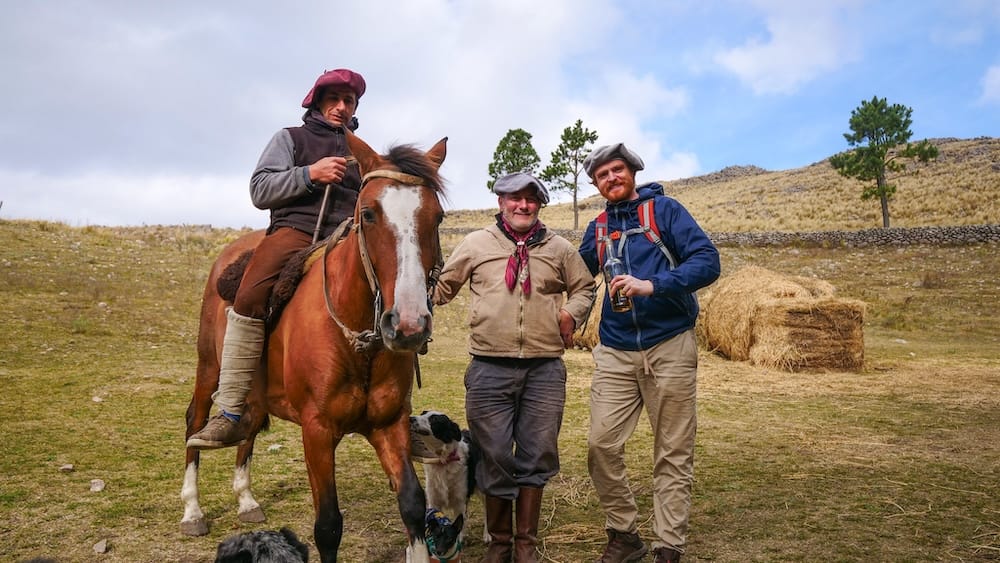 Sierras Chicas gaucho horse trek with Samuel Jeffery standing beside local riders, a saddled horse, working dogs, hay bales, and rugged mountain pasture during Project 23 Argentina fieldwork in Córdoba.
