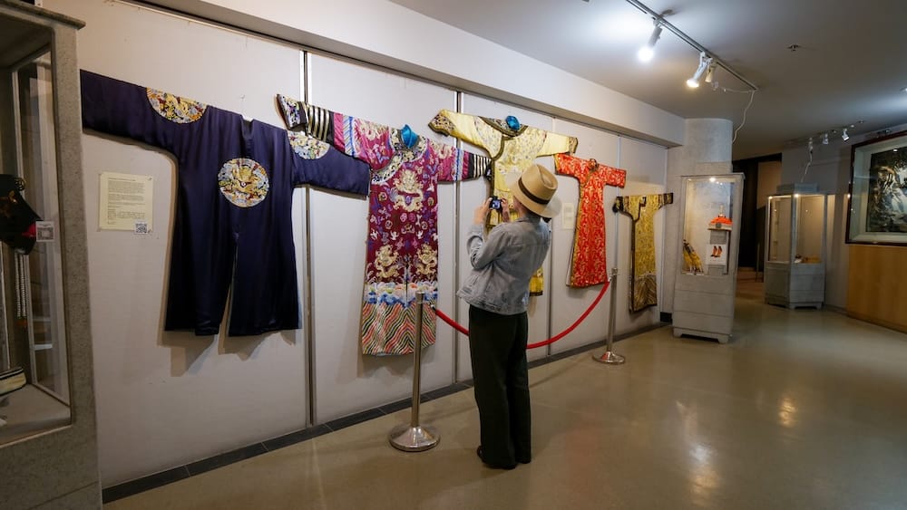 Audrey Bergner photographs a row of embroidered silk robes in navy, magenta, yellow, and red hanging on display in the Chinese Artifacts Museum, Calgary Chinese Cultural Centre, Alberta, Canada.