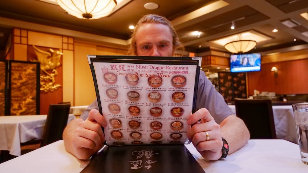 Reviewing the Evening Dim Sum Menu at Silver Dragon Restaurant, Chinatown, Calgary, Alberta, Canada Samuel Jeffery holding open Silver Dragon's laminated evening dim sum menu inside the restaurant's dining room in Chinatown, Calgary, Alberta, Canada, with photos of dumpling dishes and printed prices visible.