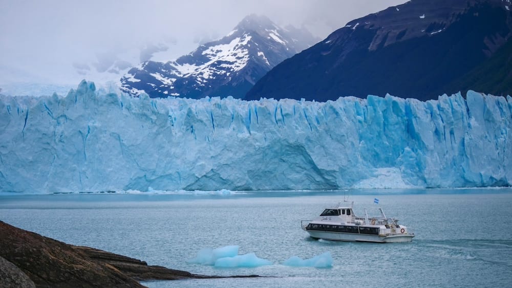 Barco de turismo Santa Fe frente a la pared del Glaciar Perito Moreno, El Calafate, Santa Cruz, Argentina El pequeño barco de turismo Santa Fe, con bandera argentina, navega junto a bloques de hielo flotante debajo de la imponente pared de hielo azul y blanco del Glaciar Perito Moreno, con montañas oscuras detrás, cerca de El Calafate, Santa Cruz, Patagonia argentina.