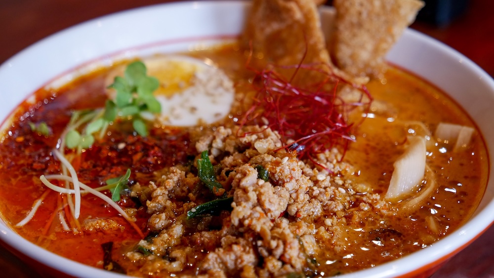 Primer plano de un plato de Enma ramen picante en Noboribetsu Onsen, Hokkaido, Japón, con caldo rojo intenso, carne de cerdo picada, medio huevo pasado por agua, brotes de bambú, hojas verdes y una guarnición crocante.