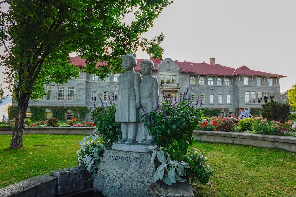 St. Eugene Golf Resort & Casino near Cranbrook, British Columbia, showing the Kamnin Tik memorial sculpture of two children in the garden, set in front of the historic Mission Building, honoring memory, reflection, and Indigenous history tied to the site.