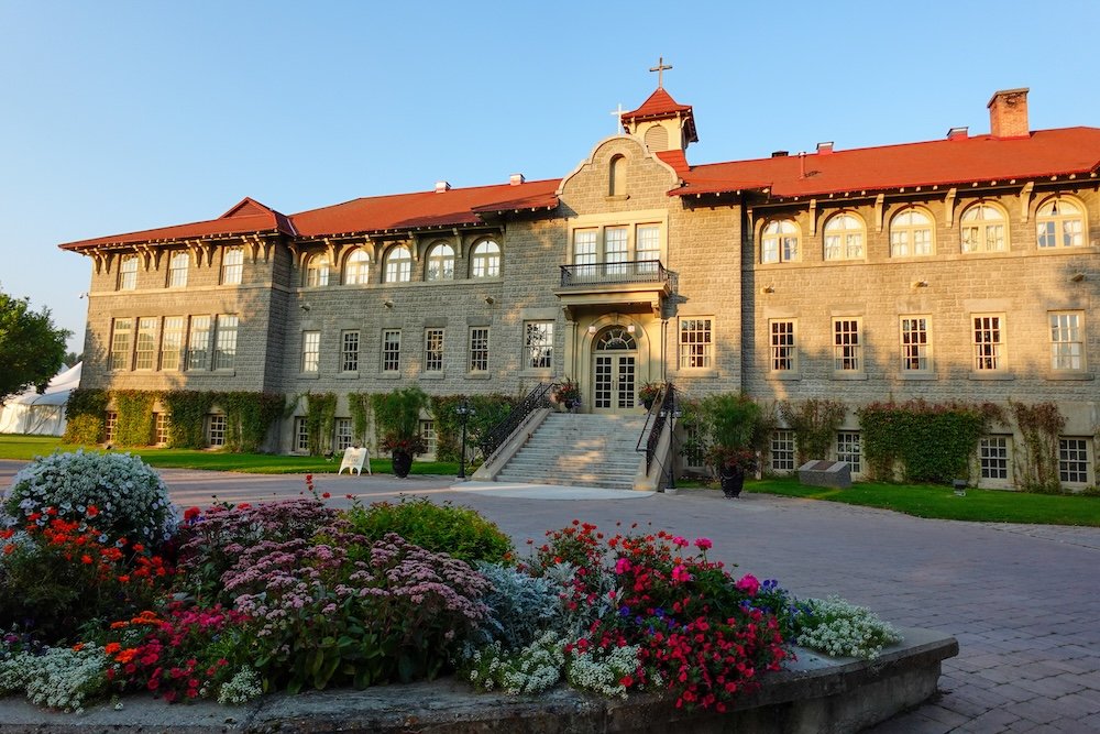 St. Eugene Golf Resort & Casino seen from the landscaped garden at golden hour, with the historic stone Mission Building glowing in warm sunlight, framed by colorful flower beds and manicured grounds near Cranbrook, British Columbia.