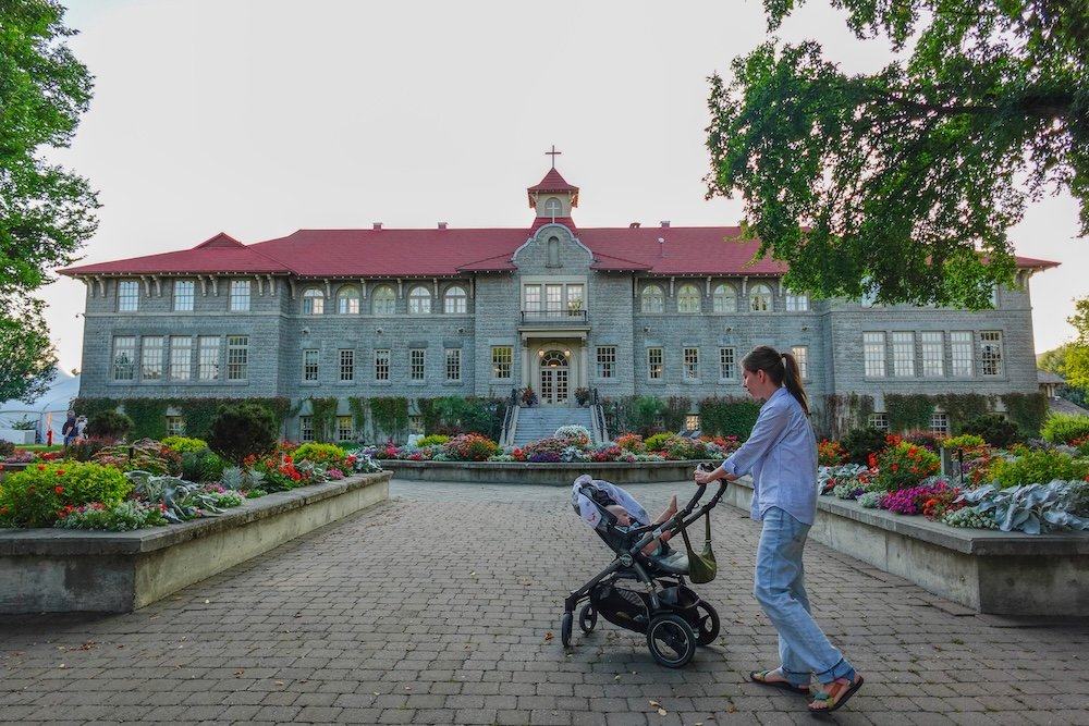 St. Eugene Mission near Cranbrook, British Columbia, featuring the historic stone building and landscaped gardens as Audrey Bergner strolls with baby Aurelia in a stroller, highlighting this peaceful heritage site as a scenic and family-friendly day trip option.
