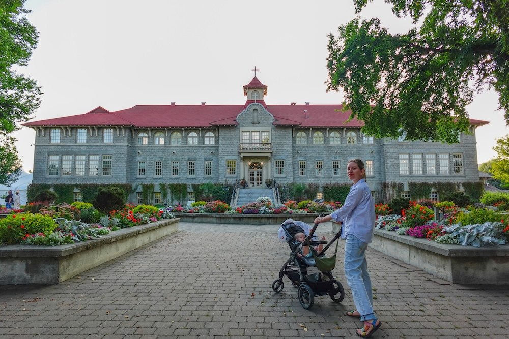 Audrey and Aurelia visiting St. Eugene Mission Resort Audrey Bergner of That Backpacker strolls with baby Aurelia in a stroller through the gardens of St. Eugene Mission Resort, a historic stone-built hotel with a red roof near Cranbrook, framed by colourful flowerbeds and soft evening light.