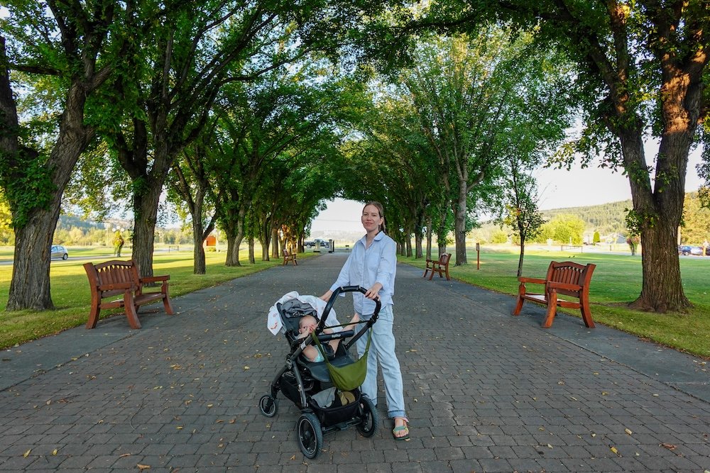 St. Eugene Mission near Cranbrook, BC, with Audrey Bergner walking along the tree-lined pathway while pushing baby Aurelia in a stroller, capturing a calm, family-friendly moment during an easy and relaxing day trip in the Kootenays.