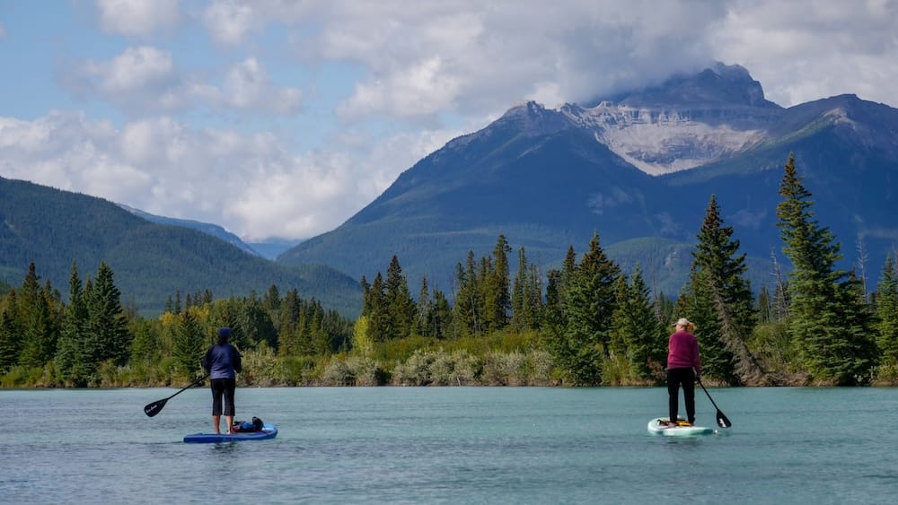 Dos personas haciendo stand-up paddle en el agua turquesa del río Bow cerca de Banff, Alberta, Canadá, con un denso bosque de pinos y una cadena montañosa con manchas de nieve de fondo.