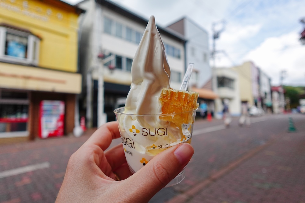 Audrey Bergner sosteniendo un vaso transparente de SUGI con helado soft cremoso y un trozo de panal de miel dorado en una calle de Noboribetsu Onsen, Hokkaido, Japón, con locales de colores y una calle adoquinada desenfocados al fondo.