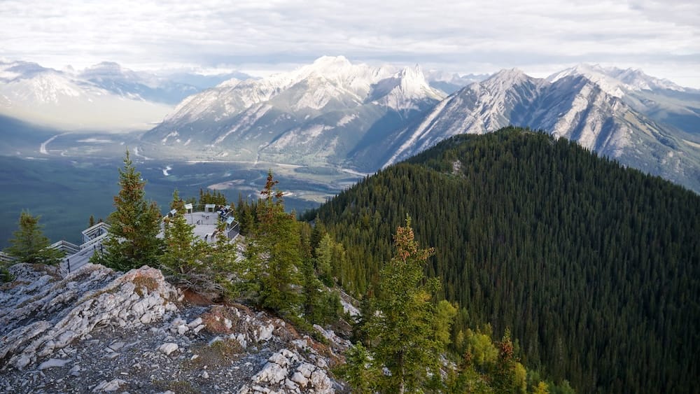 Vista desde la pasarela de Sulphur Mountain sobre Banff, Alberta, Canadá, con plataformas de la cumbre, cresta boscosa y Bow Valley Vista desde la pasarela de Sulphur Mountain cerca de la parte superior de la Banff Gondola sobre Banff, Alberta, Canadá, con terreno rocoso en primer plano, plataformas de observación debajo, crestas boscosas y amplias montañas sobre Bow Valley.