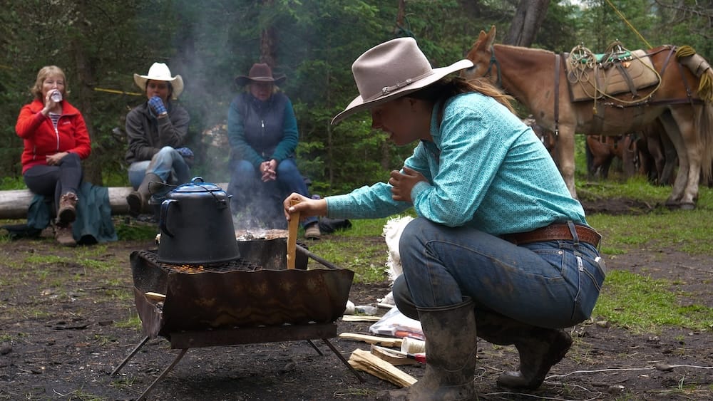 Un guía de Banff Trail Riders prepara hamburguesas a la parrilla y café sobre una cocina de campamento durante una parada para almorzar a caballo en el Parque Nacional Banff, Alberta, Canadá, con jinetes y caballos ensillados cerca.