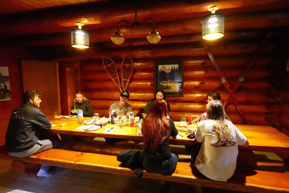 Guests eat breakfast around a long wooden table inside Sundance Lodge in Banff National Park, Alberta, Canada, with log walls, warm pendant lights, snowshoes and crossed skis decorating the room.