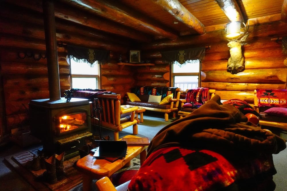 Sala junto al fuego en Sundance Lodge, Parque Nacional Banff, Alberta, Canadá, con asientos rústicos de cabaña Sala acogedora dentro de Sundance Lodge, en el Parque Nacional Banff, Alberta, Canadá, con estufa a leña encendida, paredes de troncos, asientos rústicos, mantas estampadas e iluminación cálida.