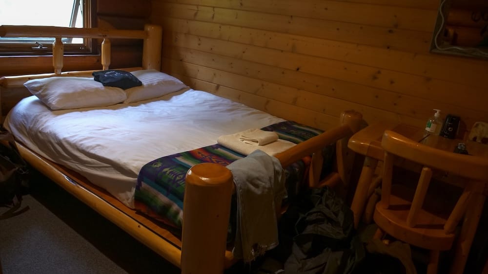 Habitación de Sundance Lodge en el Parque Nacional Banff, Alberta, Canadá, con cama de troncos e interior rústico Habitación de huéspedes dentro de Sundance Lodge, en el Parque Nacional Banff, Alberta, Canadá, con gran cama de troncos, ropa de cama blanca, paredes de madera, luz de ventana y muebles rústicos.