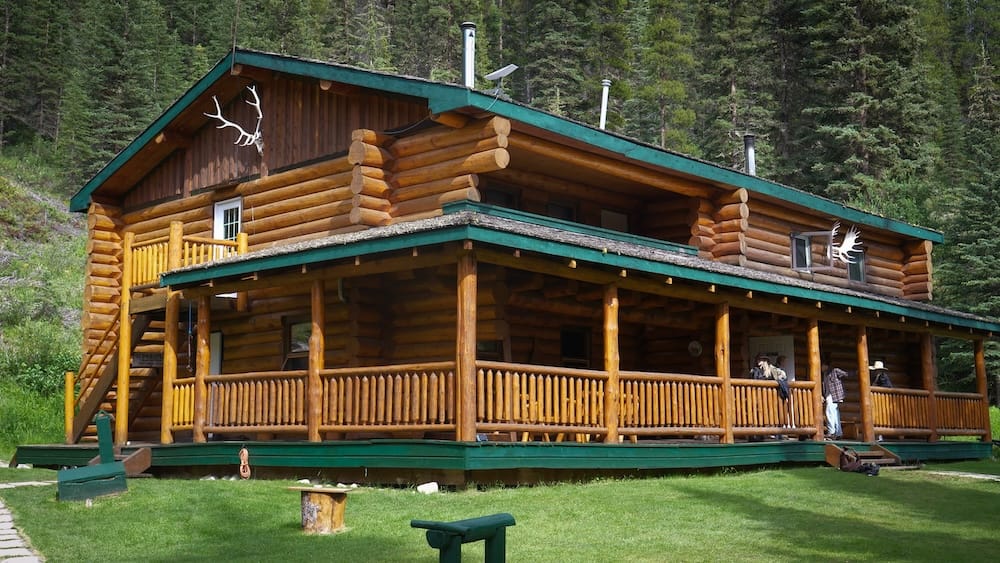 Sundance Lodge, una gran cabaña de troncos rodeada de bosque de coníferas en el Parque Nacional Banff, Alberta, Canadá, con galería perimetral y algunos huéspedes afuera.