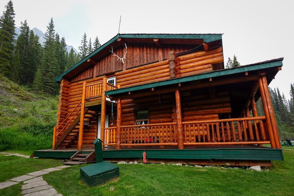 Sundance Lodge log cabin accommodation in Banff National Park, Alberta, Canada on the six-day horseback trip Sundance Lodge in Banff National Park, Alberta, Canada, shown as a large rustic log cabin with a covered porch, exterior staircase, forested slopes and mountain peaks partly visible behind the property.