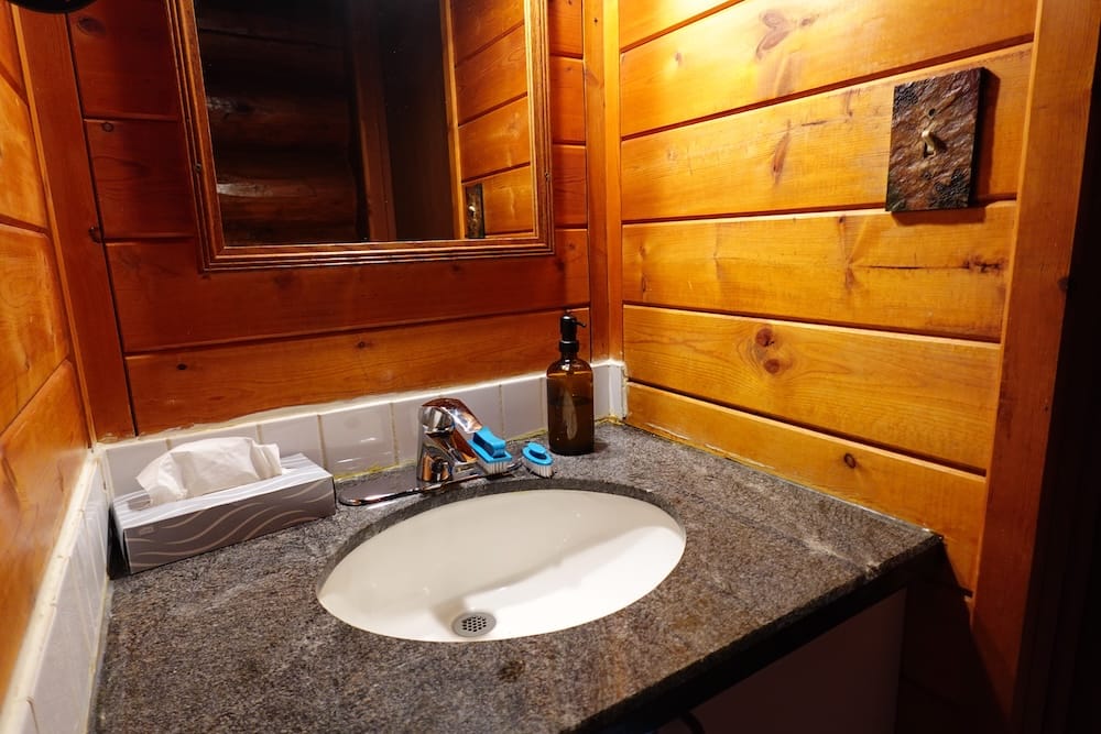 Baño interior de Sundance Lodge en el Parque Nacional Banff, Alberta, Canadá, con paredes de madera rústica y zona de lavamanos Baño interior de Sundance Lodge, en el Parque Nacional Banff, Alberta, Canadá, con mesada de piedra, pileta blanca, espejo, paredes revestidas en madera y terminaciones de estilo rústico.