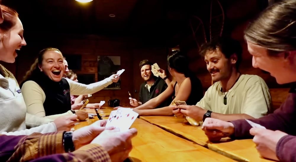Partida de cartas en Sundance Lodge, Parque Nacional Banff, Alberta, Canadá, con el grupo de jinetes alrededor de la mesa comunitaria Un grupo de jinetes riéndose y jugando a las cartas alrededor de la gran mesa de madera dentro de Sundance Lodge, en el Parque Nacional Banff, Alberta, Canadá, durante una noche en el lodge de backcountry.