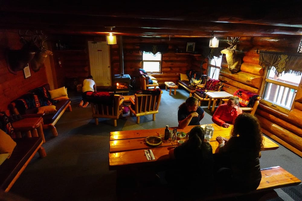 Sala común de Sundance Lodge en el Parque Nacional Banff, Alberta, Canadá, con chimenea, muebles de troncos y mesa de comedor Interior del área común de Sundance Lodge en el Parque Nacional Banff, Alberta, Canadá, con paredes de troncos, chimenea, muebles de madera y huéspedes reunidos alrededor de la gran mesa del comedor.