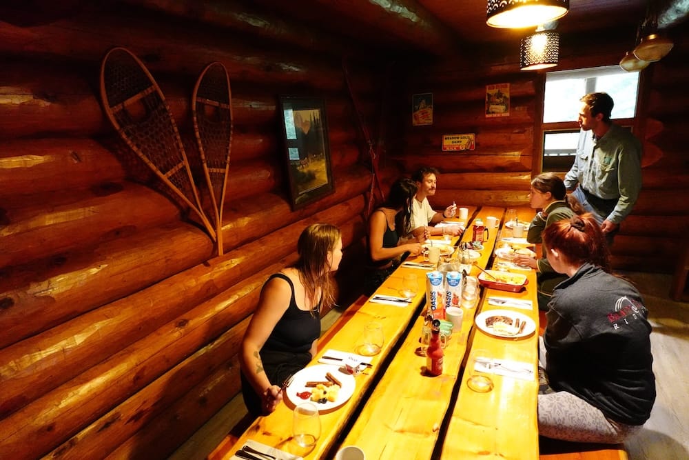 Comida comunitaria en Sundance Lodge, Parque Nacional Banff, Alberta, Canadá, dentro del comedor rústico de troncos Huéspedes compartiendo una comida alrededor de una larga mesa de madera dentro de Sundance Lodge, en el Parque Nacional Banff, Alberta, Canadá, con paredes de troncos, decoración rústica y raquetas de nieve junto al comedor.