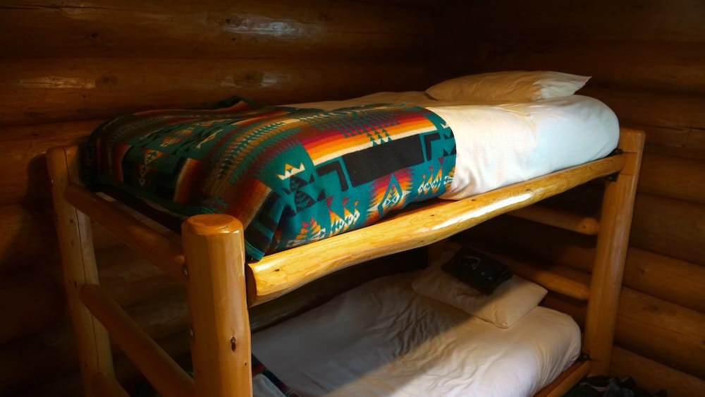 Habitación de huéspedes en Sundance Lodge, Parque Nacional Banff, Alberta, Canadá, con paredes de troncos y camas cucheta Camas cucheta rústicas dentro de una habitación de Sundance Lodge, en el Parque Nacional Banff, Alberta, Canadá, con paredes de troncos, estructuras de madera, ropa de cama blanca y una manta estampada en la cama superior.