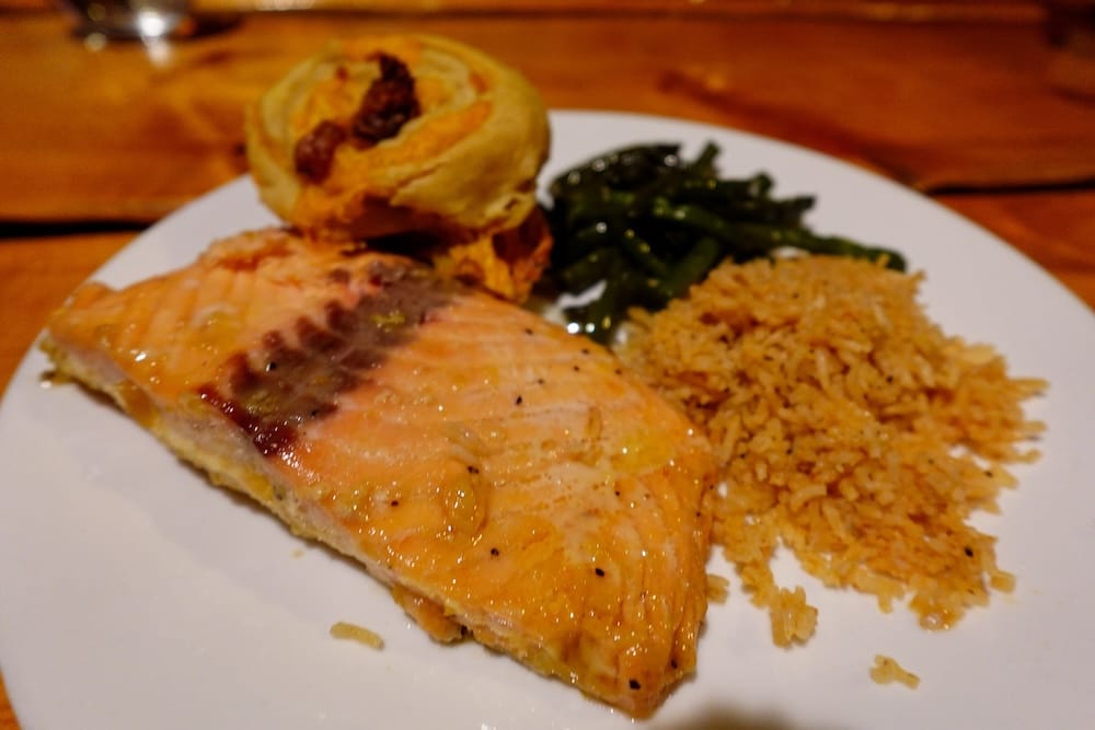 Cena de salmón en Sundance Lodge, Parque Nacional Banff, Alberta, Canadá, durante una estadía de backcountry a caballo Cena de salmón servida en Sundance Lodge, Parque Nacional Banff, Alberta, Canadá, con arroz, verduras verdes y una guarnición horneada sobre un plato blanco en la mesa comunitaria de madera.