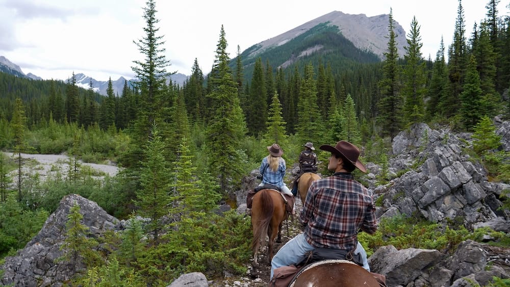 Jinetes avanzan por un sendero rocoso entre Sundance Lodge y Halfway Lodge, dentro del Parque Nacional Banff, Alberta, Canadá, rodeados de coníferas, roca expuesta y montañas.