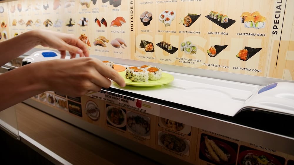 Una mano levantando un plato de rollos de sushi de una cinta transportadora, con fotos del menú y un cartel que dice "por favor espere al tren" a la vista en Point Sushi en Calgary, Alberta, Canadá.