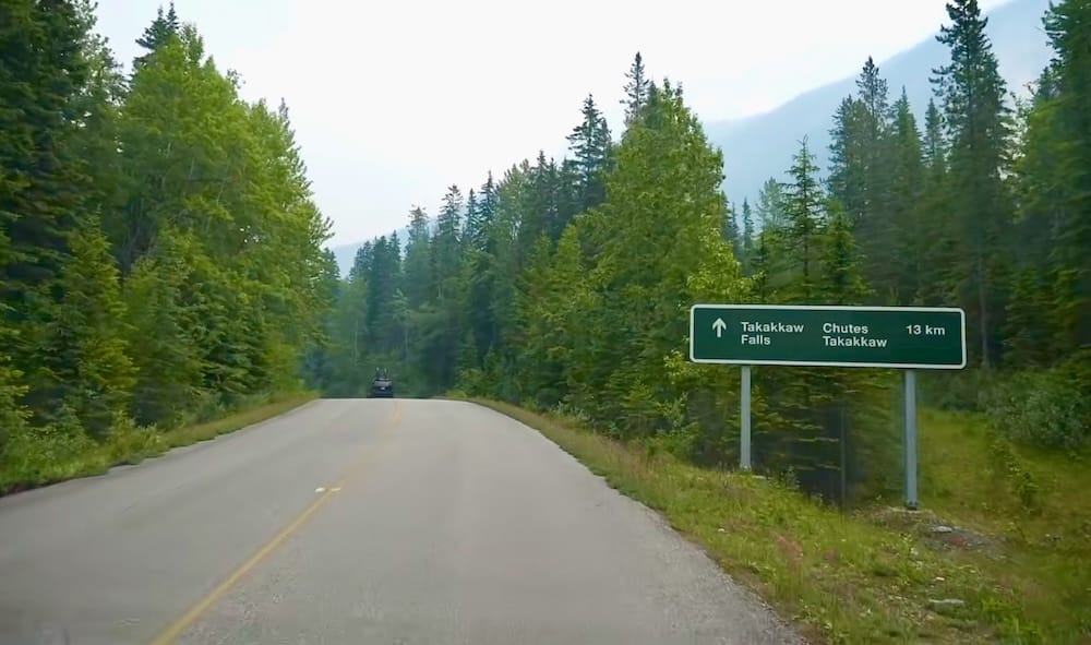 A bilingual road sign reading "Takakkaw Falls 13 km" beside Yoho Valley Road in Yoho National Park, British Columbia, with a vehicle driving ahead through dense forest toward the falls.
