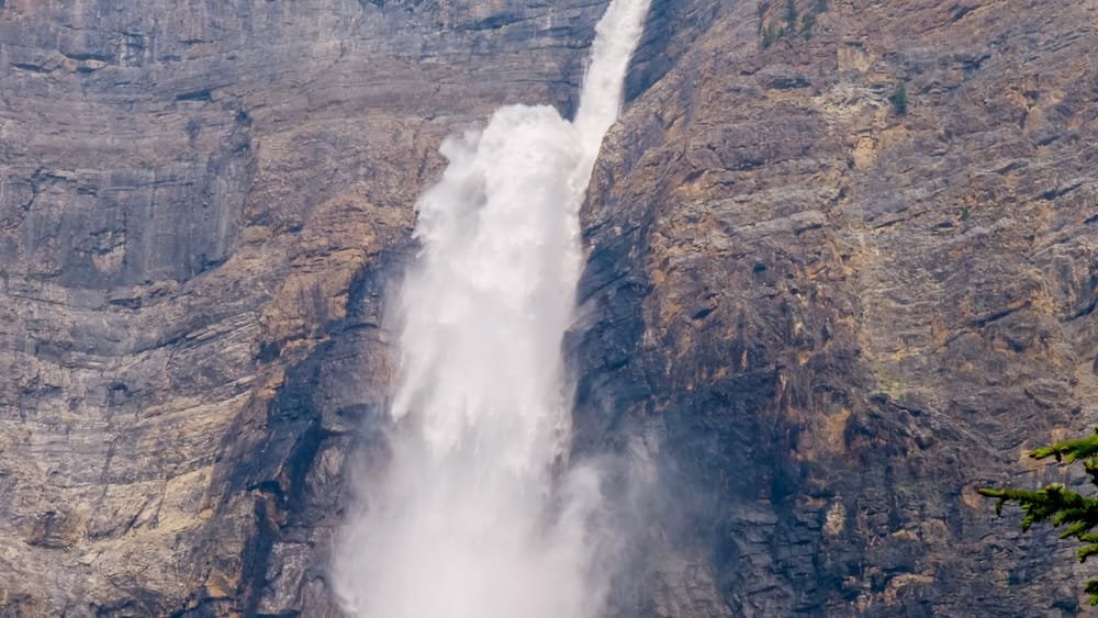 A tight close-up of Takakkaw Falls in Yoho National Park, British Columbia, showing the narrow jet of water striking the rock face partway down and exploding outward into a dense cloud of spray.